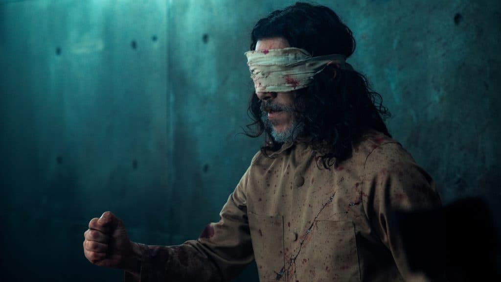 Dagin Babi el discípulo con traje marrón y los ojos vendados frente a una pared de concreto